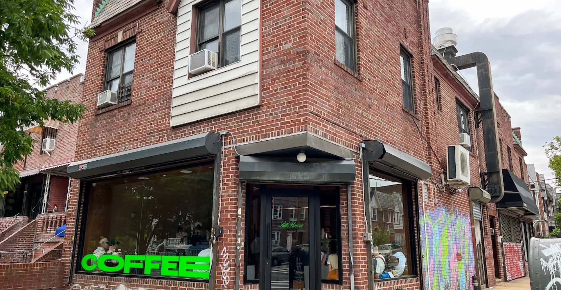 Brick exterior storefront of Little Flower Cafe. The word "coffee" is brightly painted on the bottom of the window. Graffiti covers another wall.