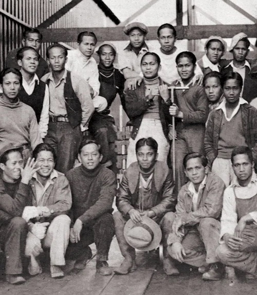 Alaskeros pose inside a cannery with six men crouching in front of fourteen others. They are dressed in pants, vests, jackets, hats, collared shirts.