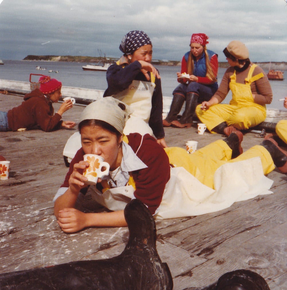 Five female cannery workers, dressed in aprons, overalls, rubber boots, and bandanas, rest on a dock by the ocean and drink out of paper cups.