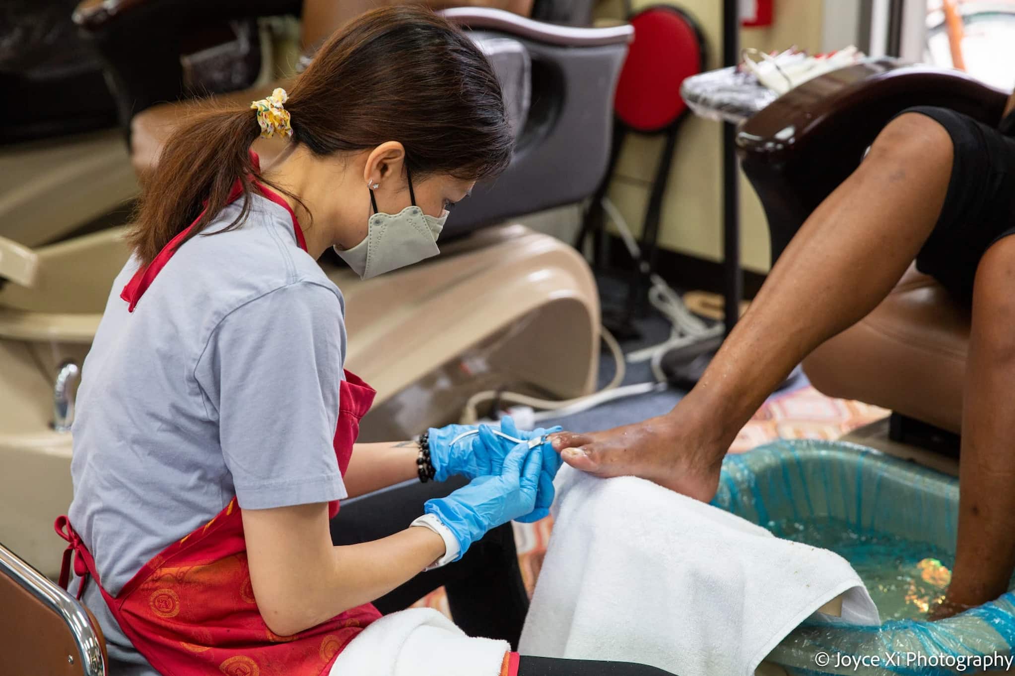 A Vietnamese female nail tech in an apron, face mask and latex gloves gives a customer a pedicure while seated in front of the customer and foot bath.