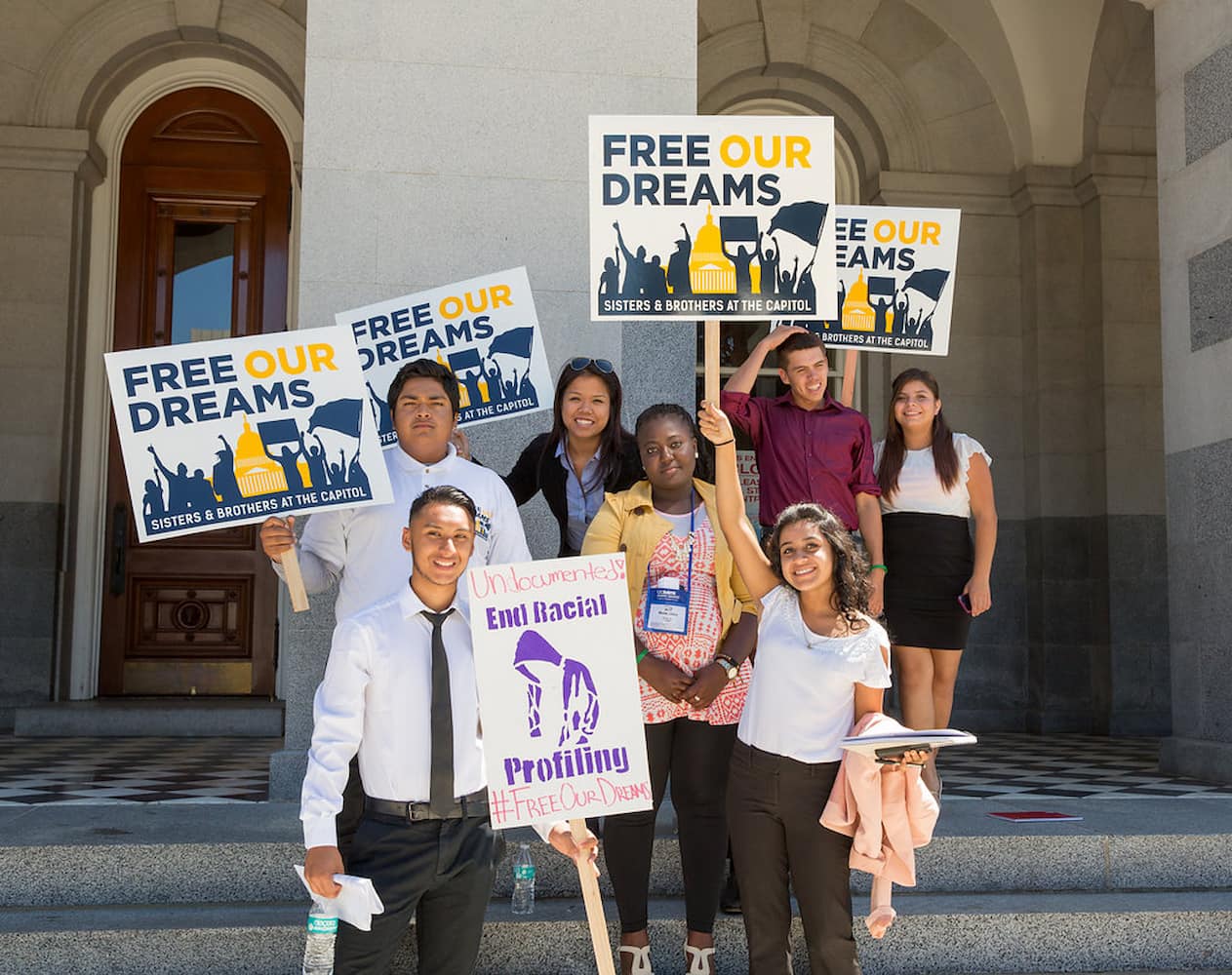 Six young adults of varying ethnicities and one woman stand at the steps outside of a building holding signs that read "Free Our Dreams."