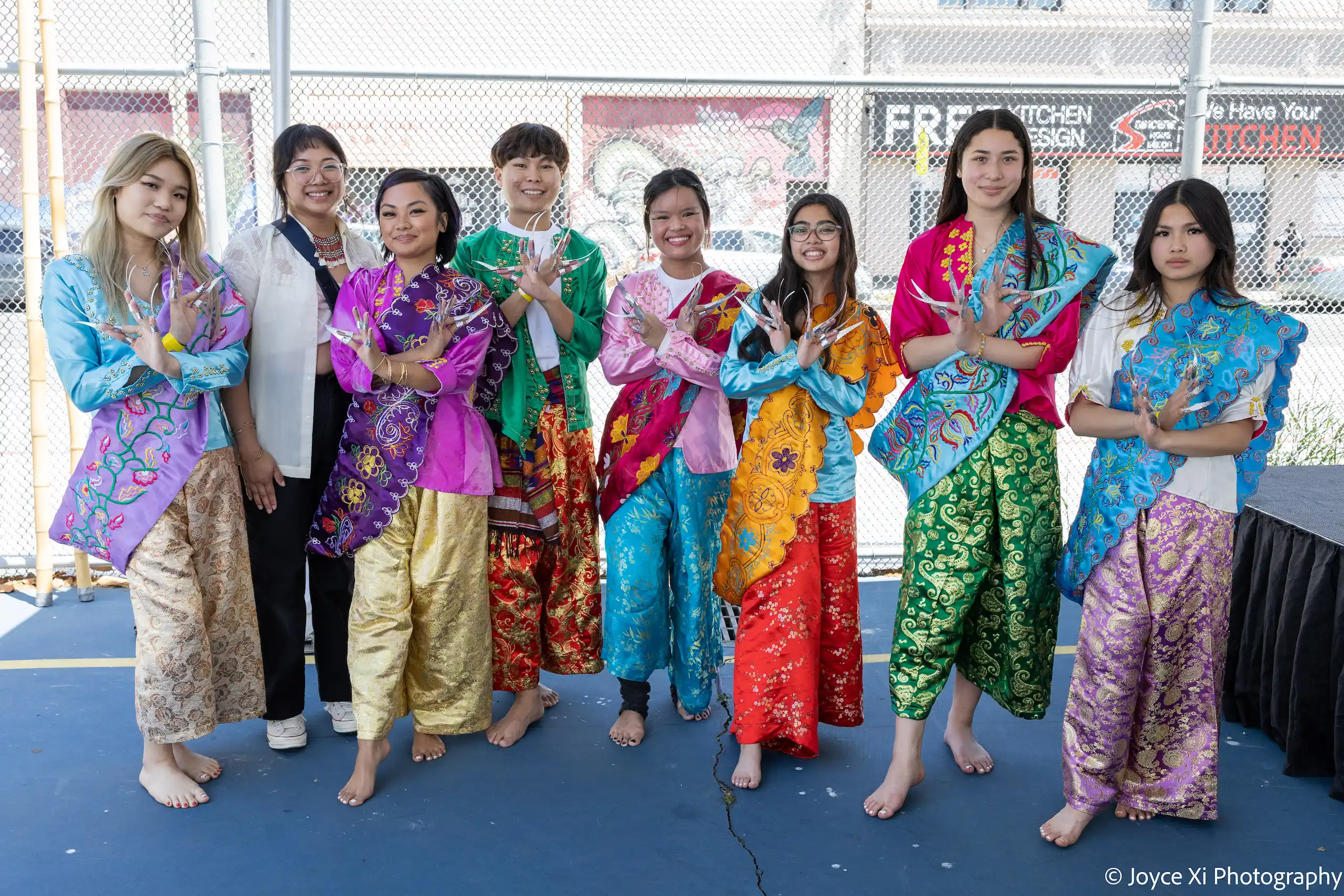 Eight young adults pose for photo near outdoor stage. Seven of them wear brightly colored garments and pose with their hands crossed in front of them.