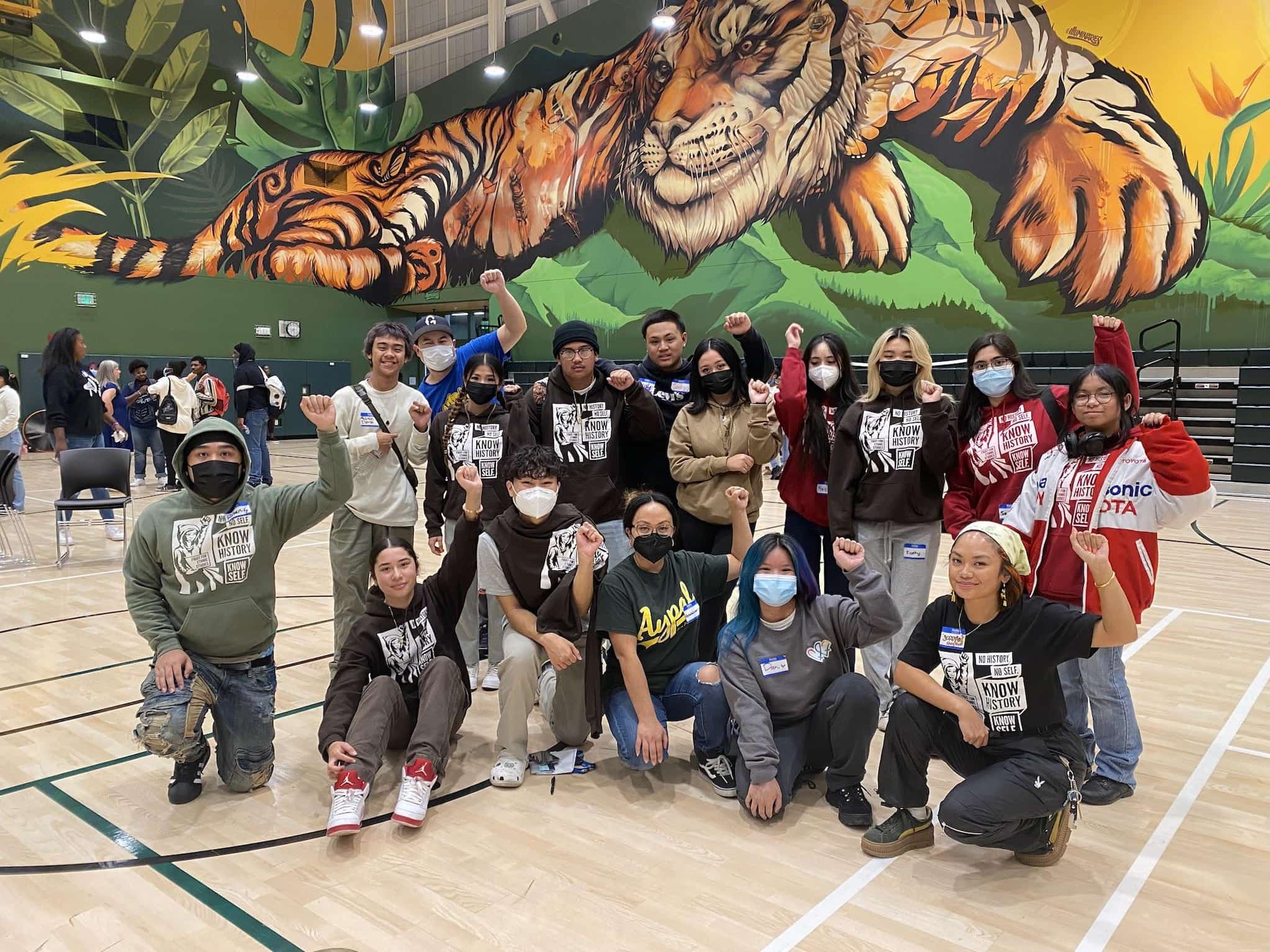 Sixteen AYPAL members pose with their fists raised in the air for a photo in a school gym. Behind them is a wall mural of a large tiger.
