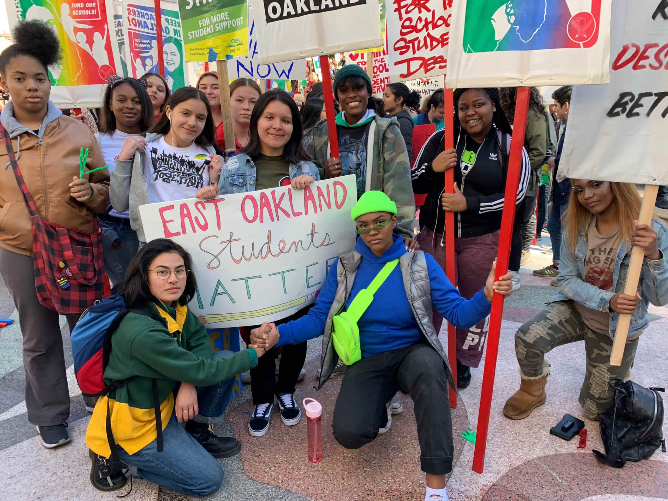 Twelve Youth Together members pose for a picture while marching in a picket line. They hold signs with red sticks expressing support for teachers.