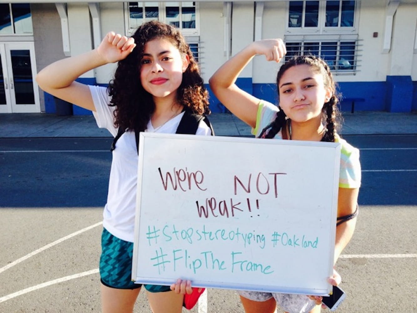 Two young women hold a whiteboard that reads "We're not weak!!" while flexing their right arms. They are standing on a blacktop in front of a school.