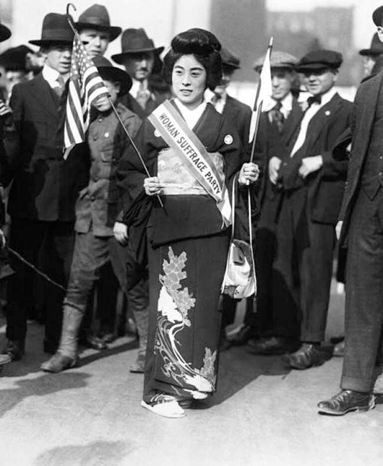 Komako Kimura, a Japanese suffragist dressed in a kimono, marches with the flags of U.S. and Japan in each hand. Men in suits crowd around her.