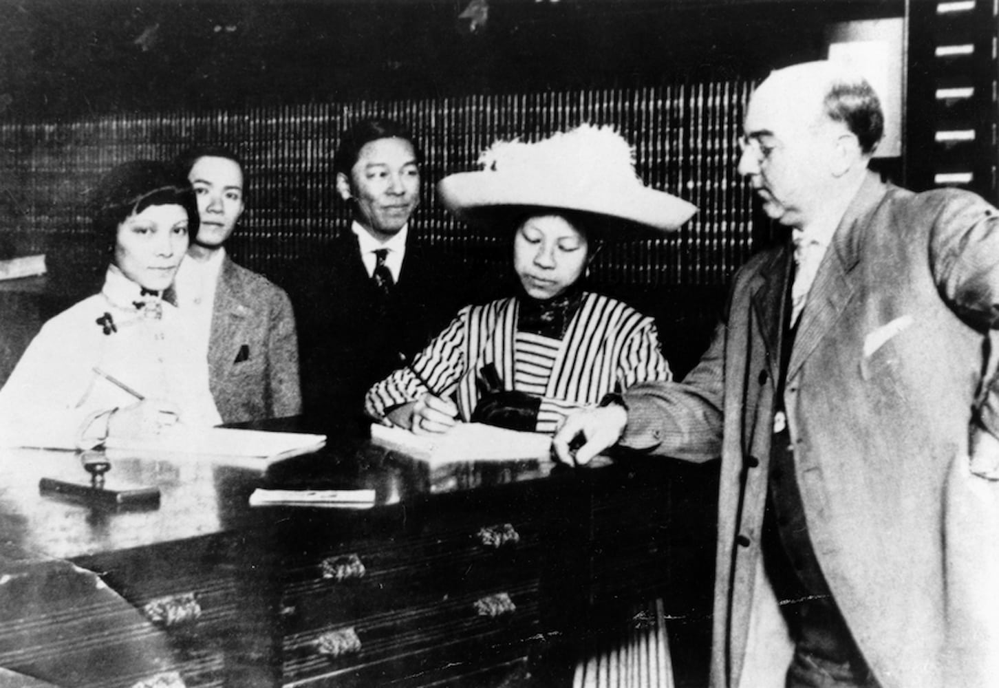 Clara Elizabeth Chan Lee, in a striped dress and large hat, registers to vote. Her husband, friend, friend's husband, and clerk stand around her.