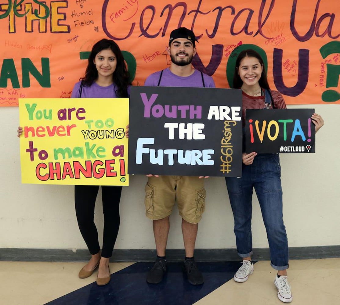 One male student stands between two female students as they pose with signs against hallway wall. Their handmade signs promote civic engagement.