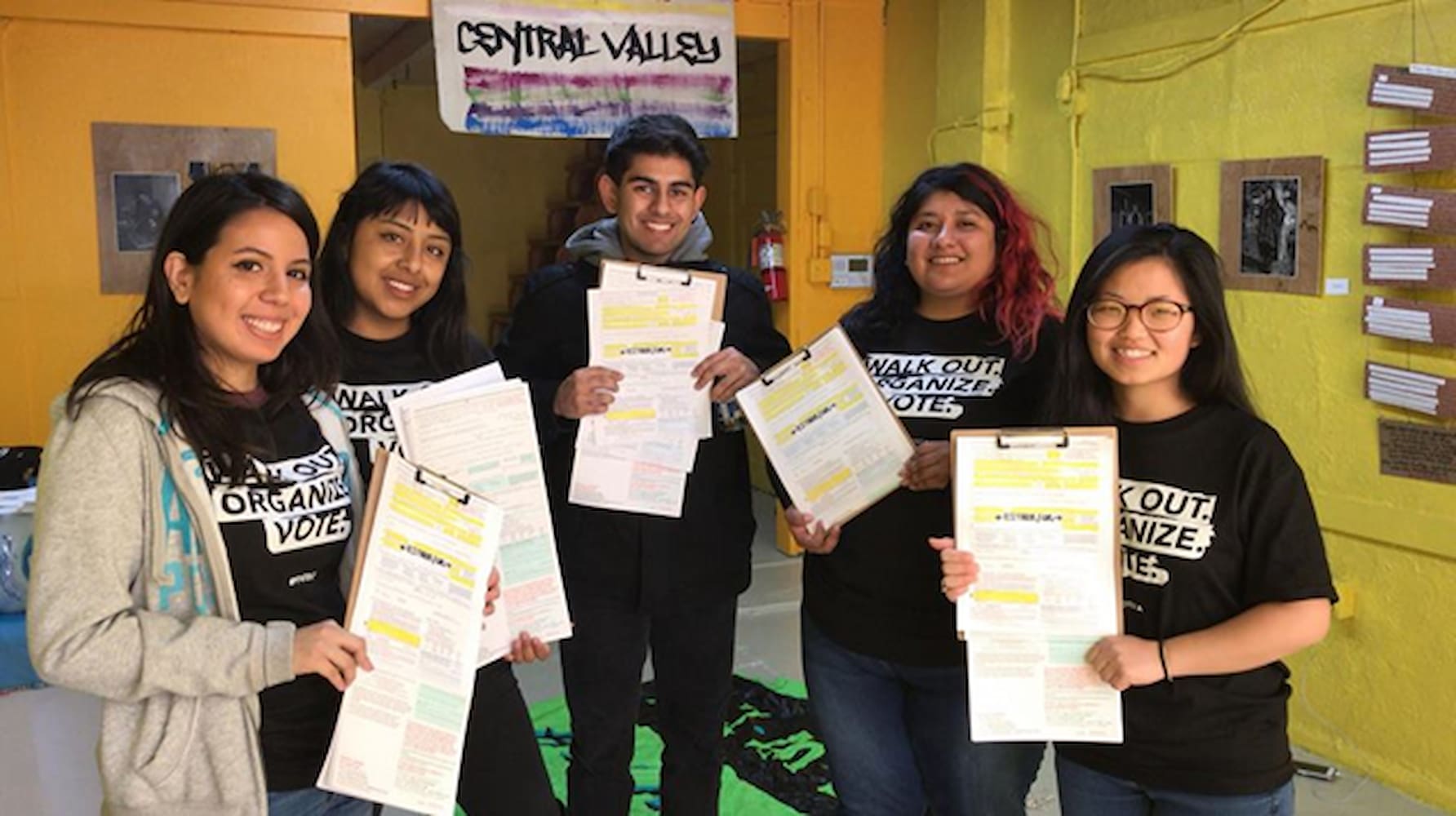 One male student stands with four female students as they pose with clipboards and papers in school hallway during voter registration drive.