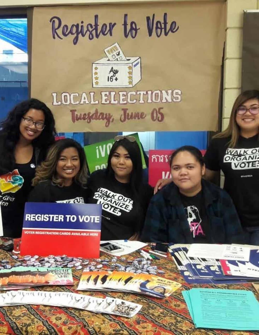 Five Khmer Girls in Action members pose for group photo behind a voter outreach table. A "Register To Vote" banner hangs behind them.
