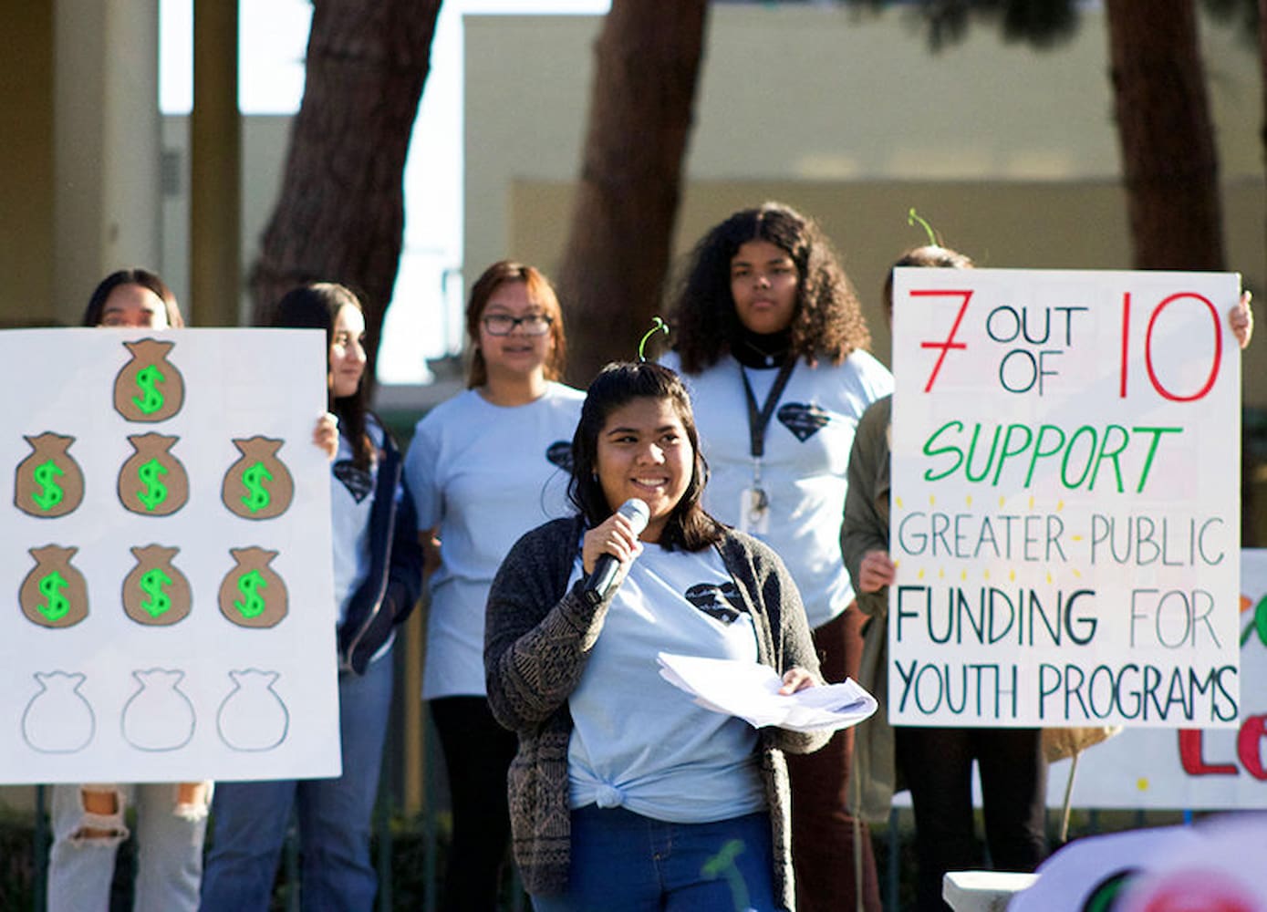A female student speaks into a microphone outdoors. Behind her are five students holding signs on funding for youth programs.