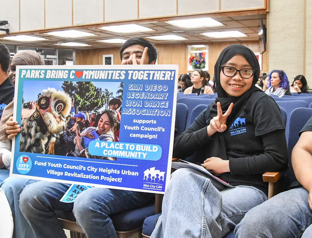 Two MCC Youth Council members, seated at hearing, pose with peace signs. On the left, a Youth Council member holds a sign that advocates for parks.