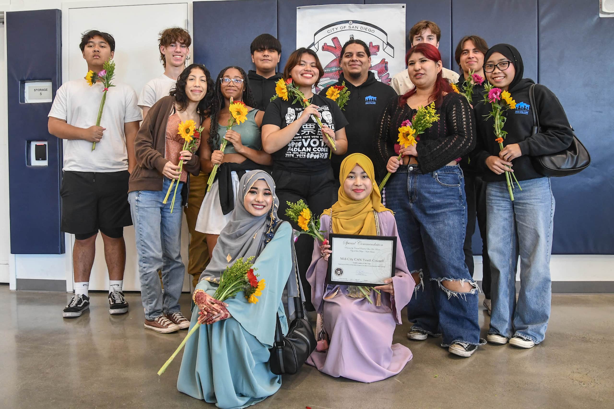 Thirteen young adults pose for indoor group photo. Two Youth Council leaders kneel at front, while others stand. Each carries a small floral bouquet.