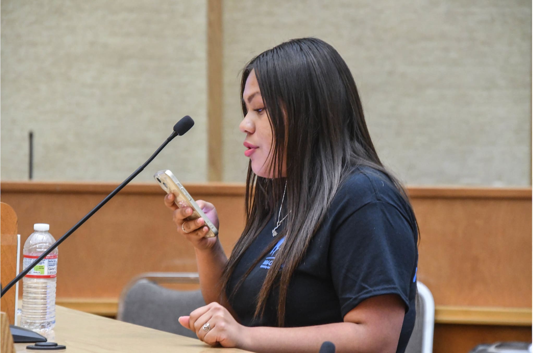 A MCC Youth Council representative, a young woman dressed in black shirt, stands in front of microphone while looking towards cell phone in her hand.