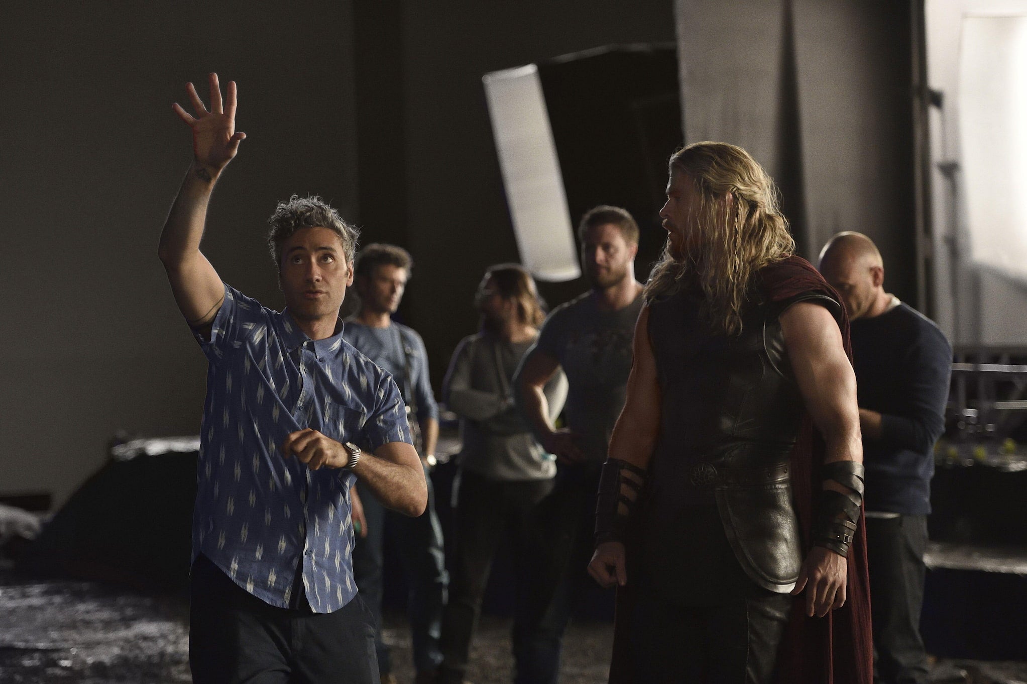 Māori filmmaker Taika Waititi raises his arm to direct Chris Hemsworth, who stands beside him in his Thor costume. Crew stand behind them on set.