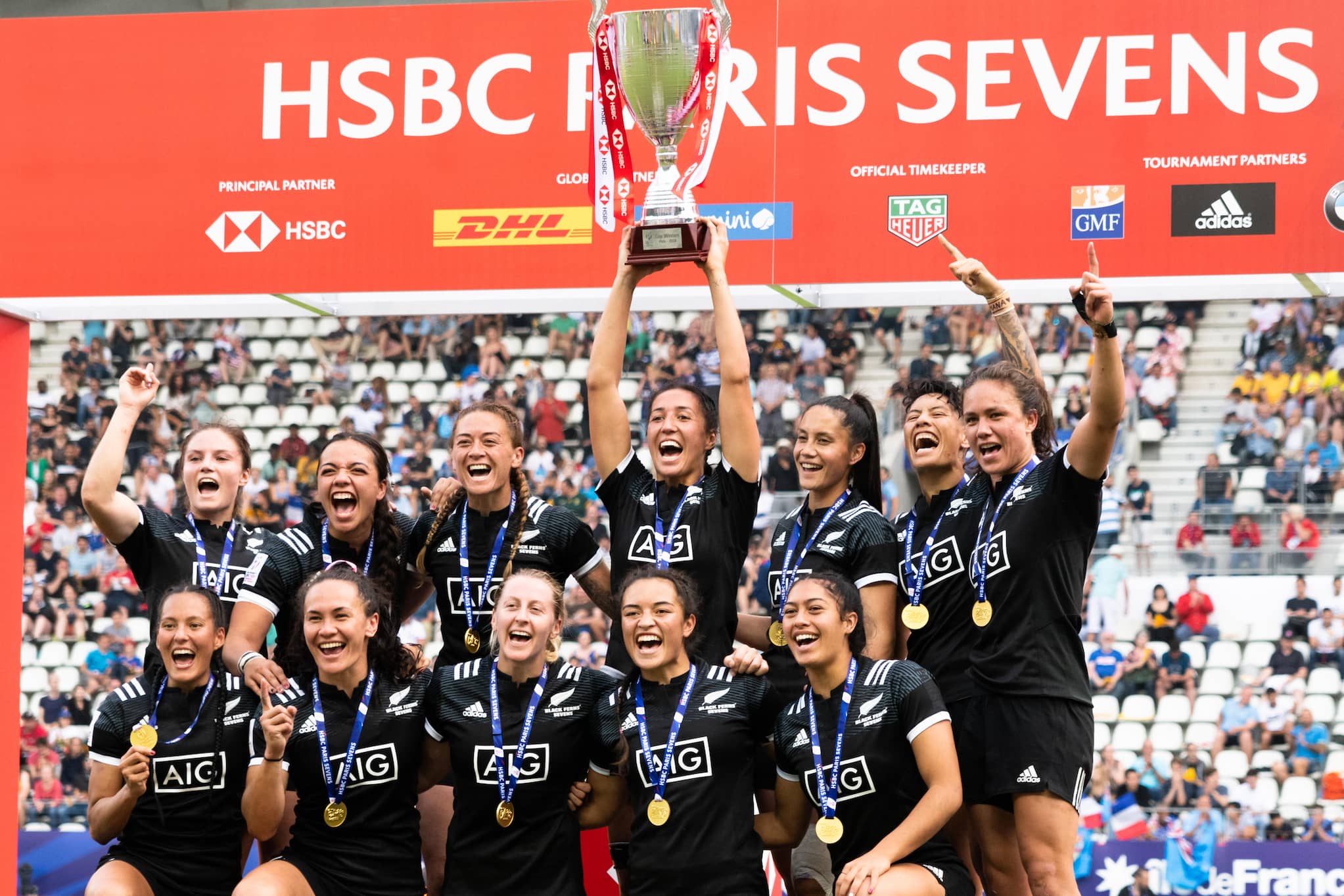Black Ferns Sevens team pose as winners of the HSBC Paris Sevens tournament. Six women kneel in front of six others with gold medals and a trophy.