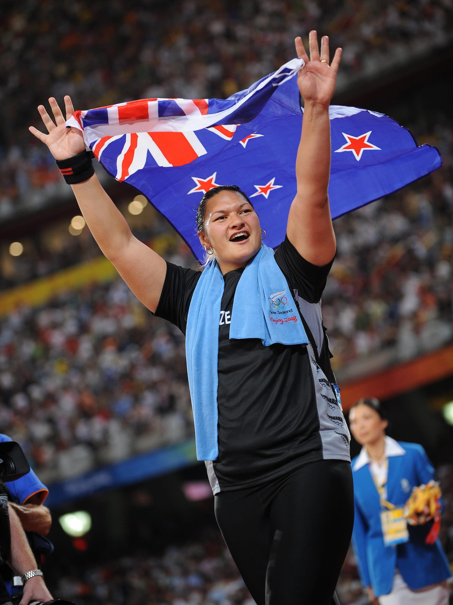 Shot putter Valerie Adams, with a towel around her neck, waves to the audience as she holds a New Zealand flag over her head at an Olympic stadium.