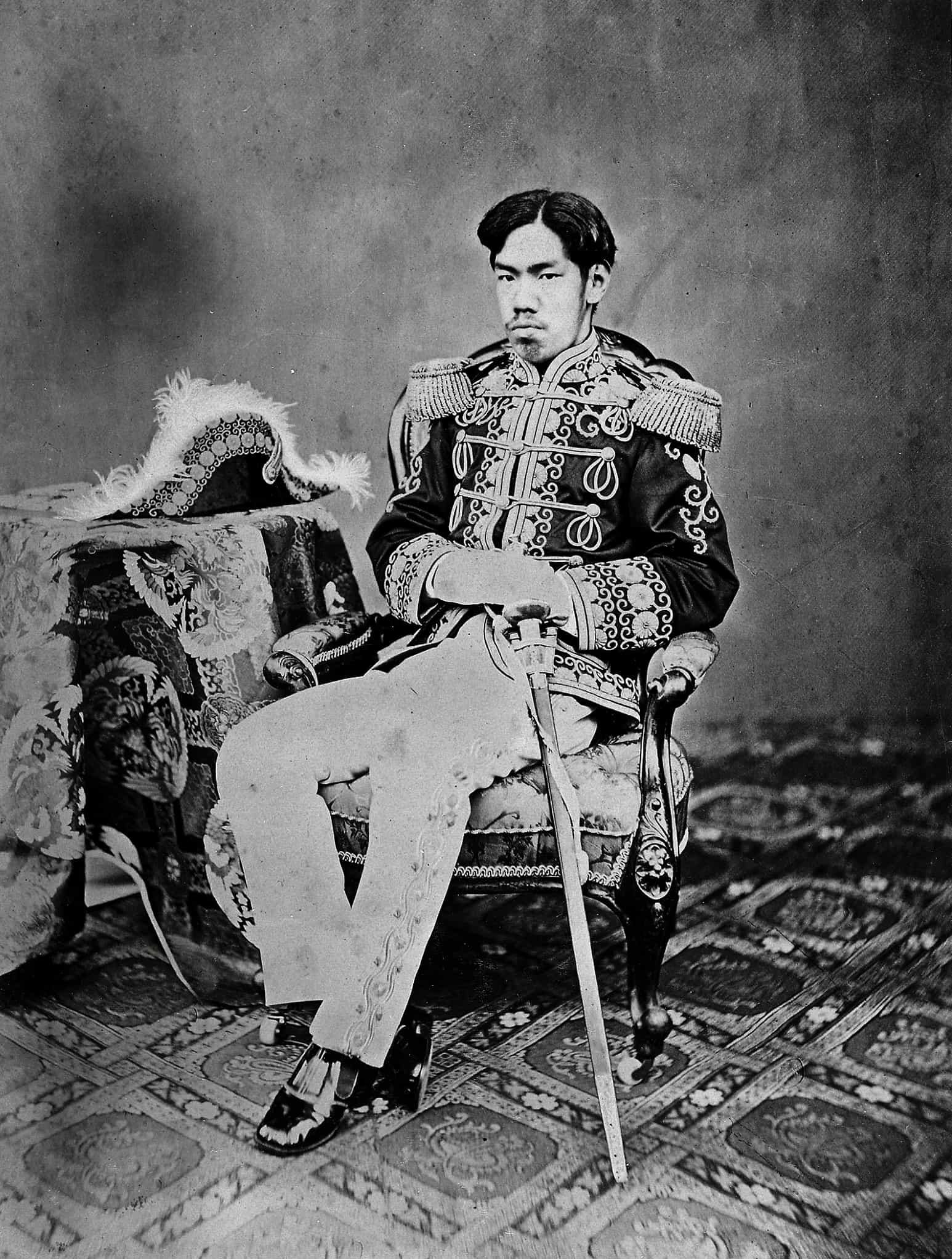 Portrait of Emperor Meiji seated in full Western-style military dress. A bicorne hat sits on a table and a sheathed sword is propped up against him.