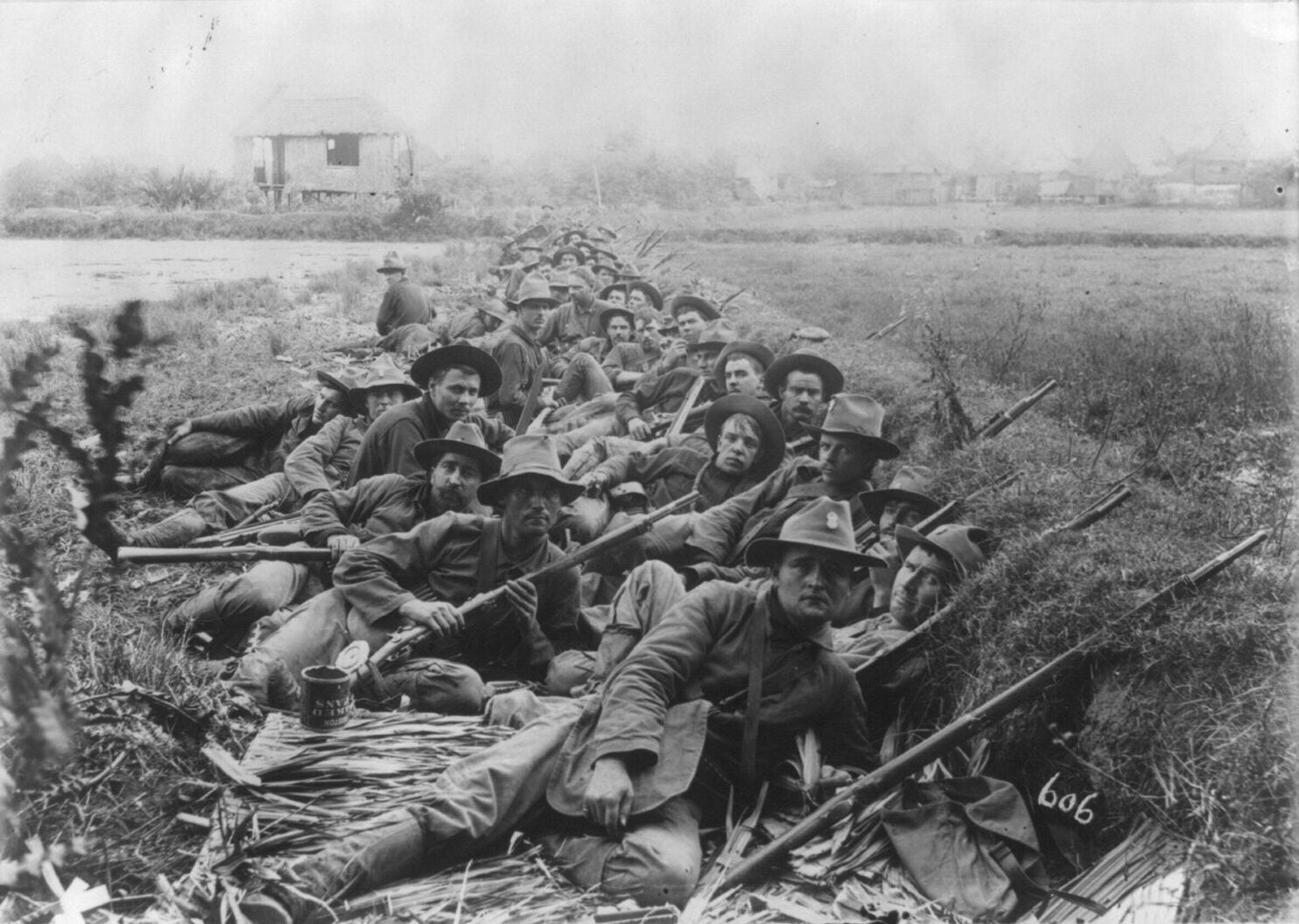 U.S. Army soldiers wearing wide brimmed hats rest in a grassy trench with their rifles leaning on the trench wall. A stilt house stands behind them.
