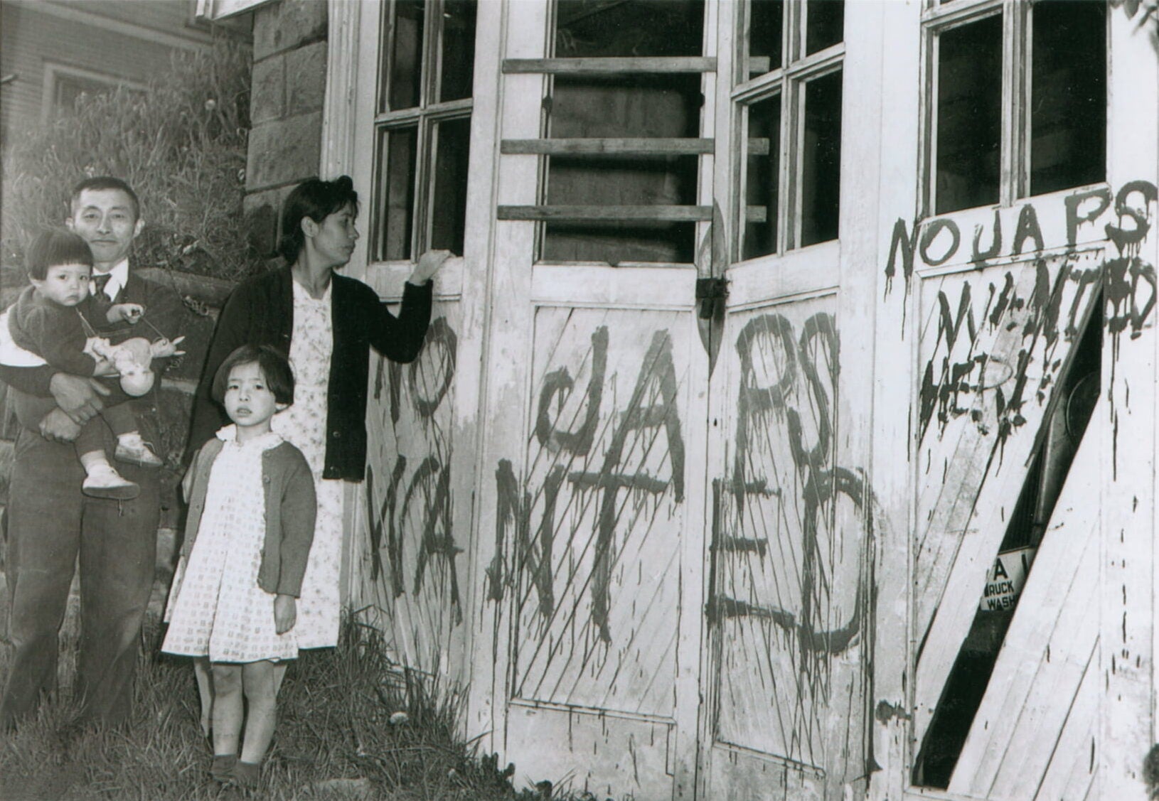 Japanese American couple and their two children stand outside their vandalized garage. Spray painted graffiti reads "No Japs Wanted" on exterior wall.