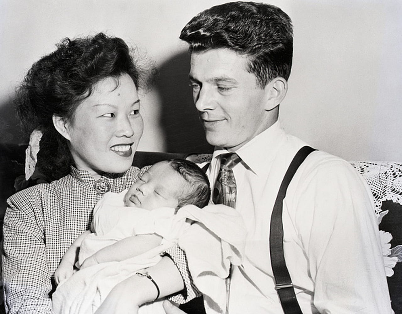 Korean military bride Yoong Soon smiles while holding her newborn daughter. She is seated next to her husband Sergeant Johnnie Morgan on a couch.