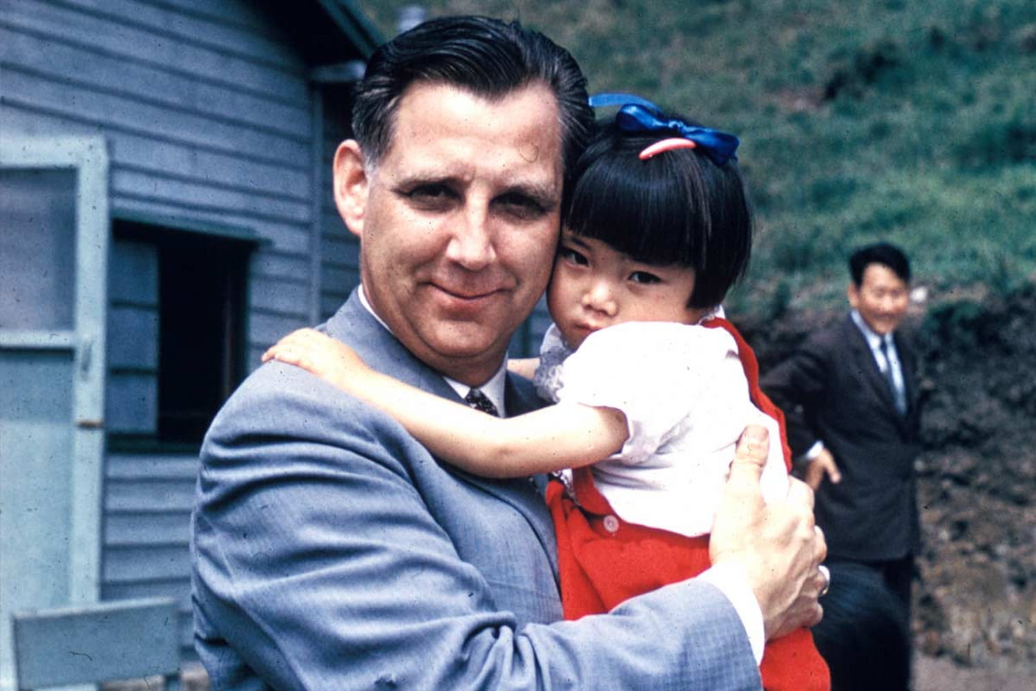 Everett Swanson, dressed in a suit, smiles at camera while holding a young South Korean war orphan, dressed in a red dress and a white shirt.