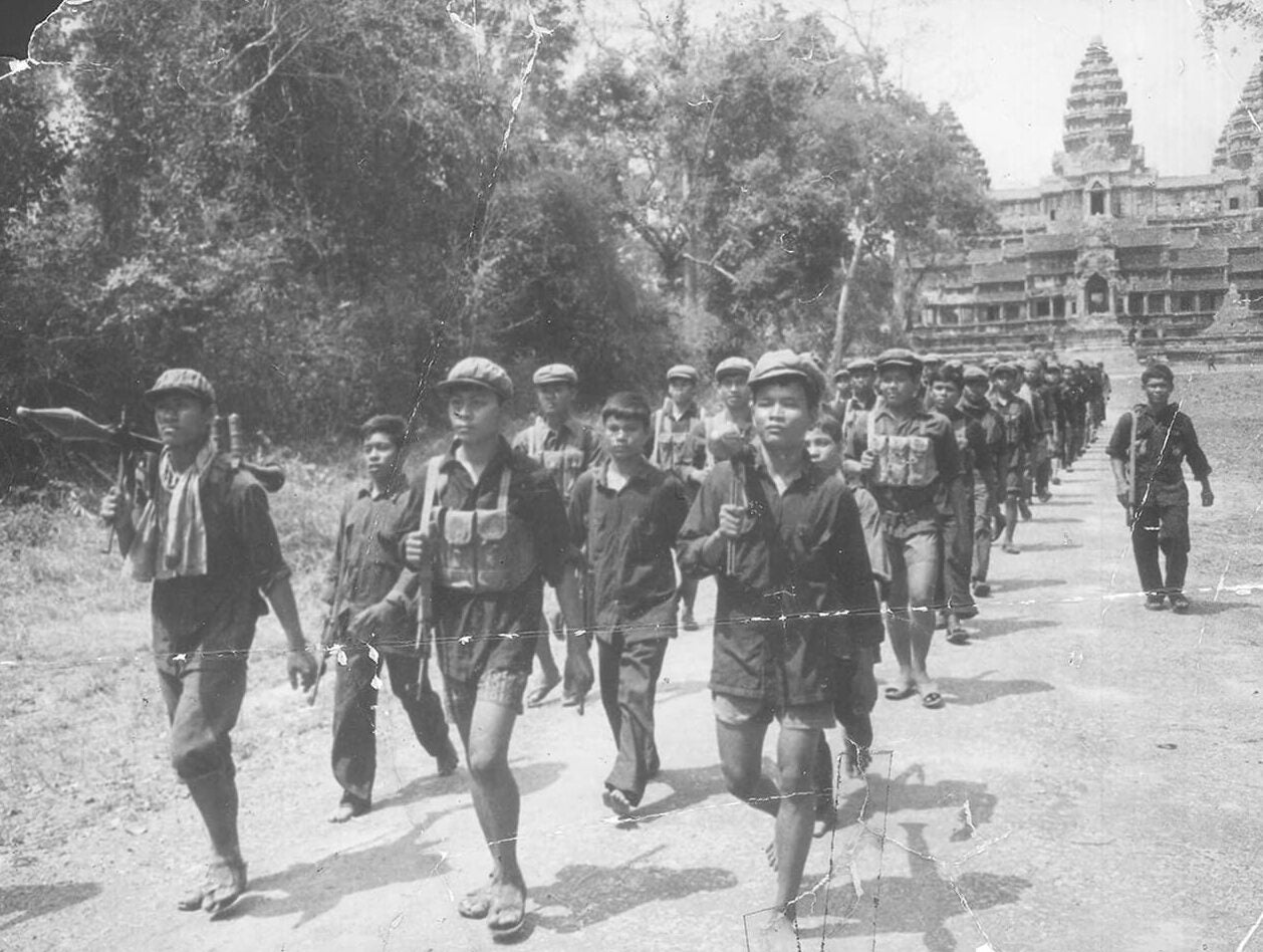 Khmer Rouge soldiers march in three lines on a dirt path with Angkor Wat behind them. They wear black shirts and shorts, sandals, and flat caps.