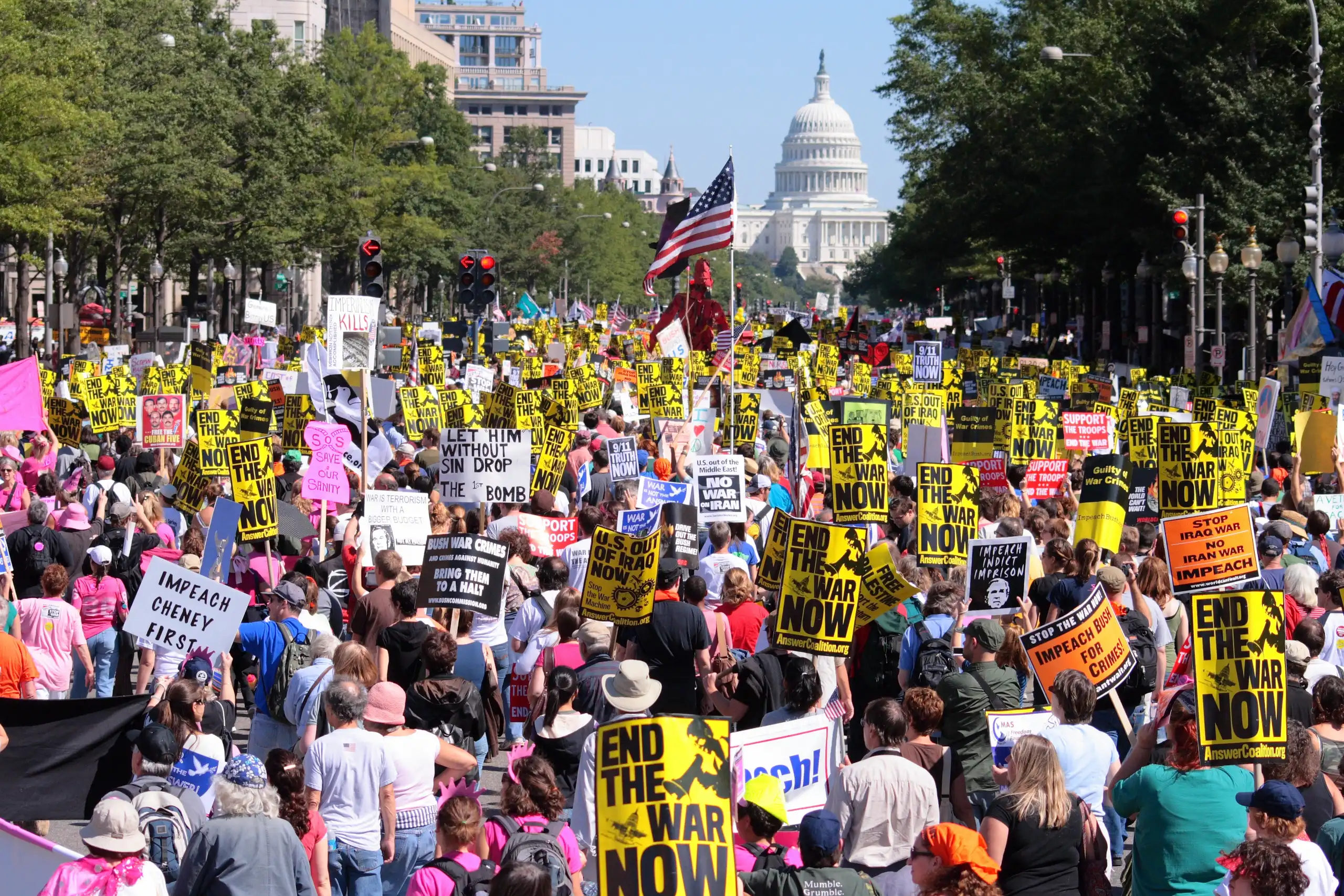 Large group of anti-war protestors march down Pennsylvania Avenue towards U.S. Capitol. They hold "End the War Now" posters, U.S. flags, and signs.