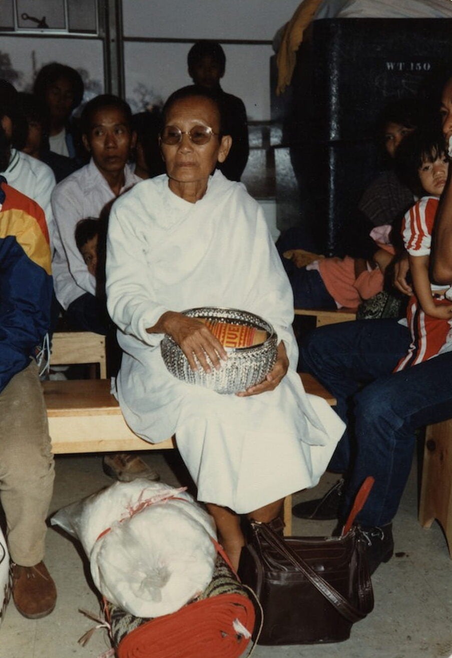 A Lao refugee, sitting on an indoor bench, carries a decorated silver bowl in her lap. A bag, mat, and purse lays at her feet.