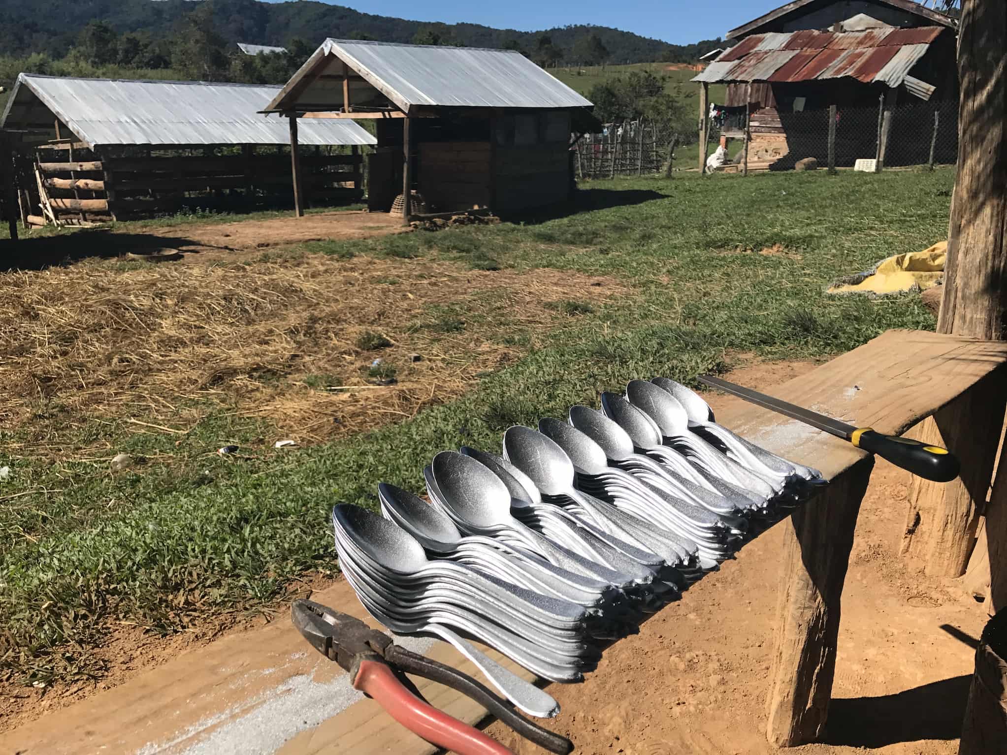 On a crude wooden bench are stacks of metal spoons between a set of pliers and a metal file. In front of the bench are three tin roofed structures.