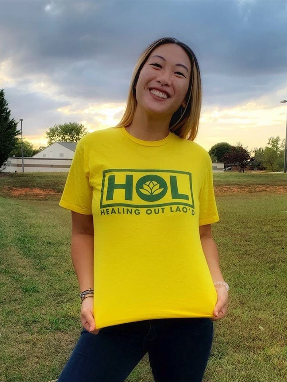 Rita Phetmixay poses with a yellow shirt in a field on a cloudy day. The shirt reads "HOL Healing Out Lao'd."