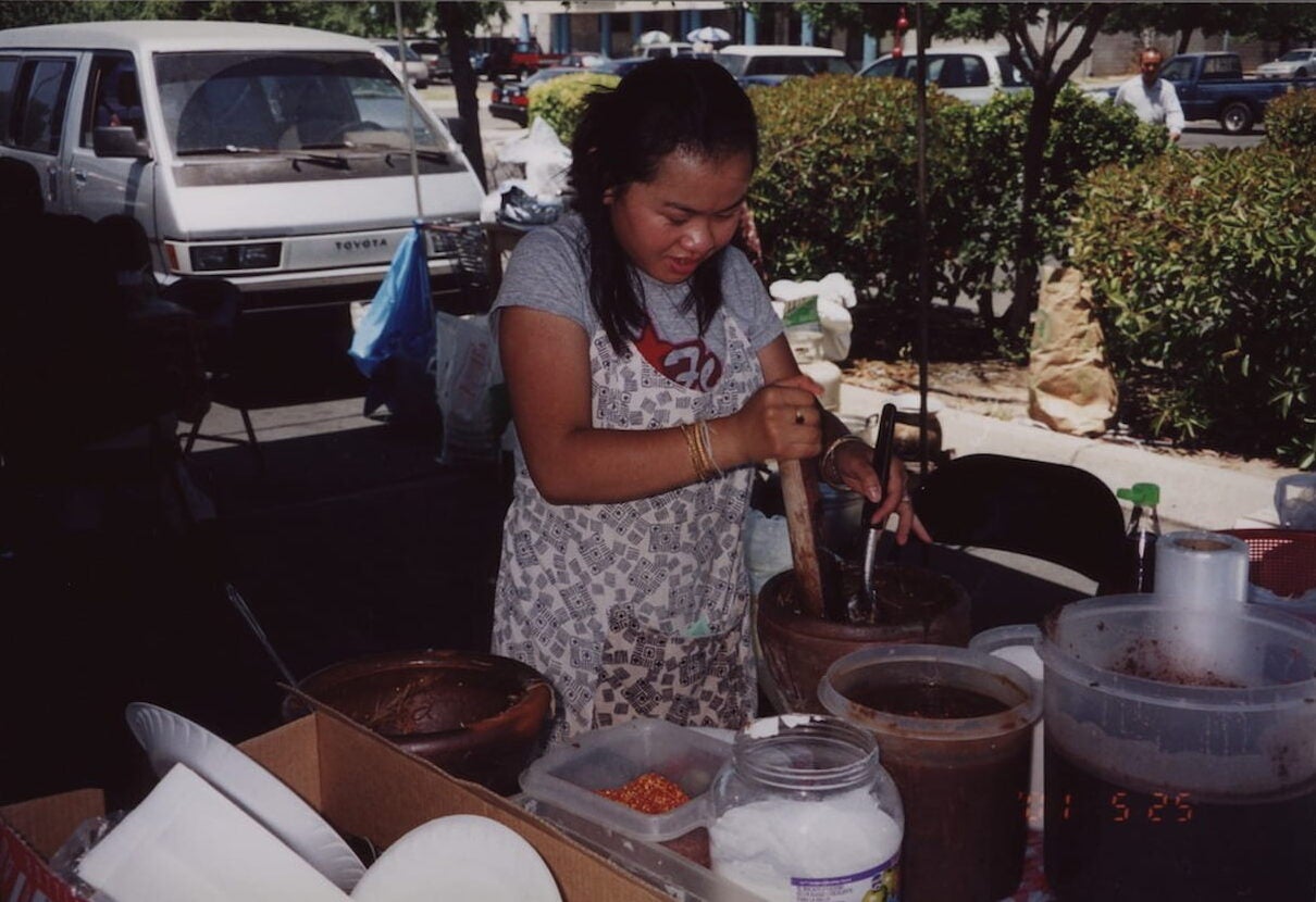 A woman in a patterned apron prepares papaya salad in an outdoor tent using a clay mortar, a wooden pestle, and a metal spoon.