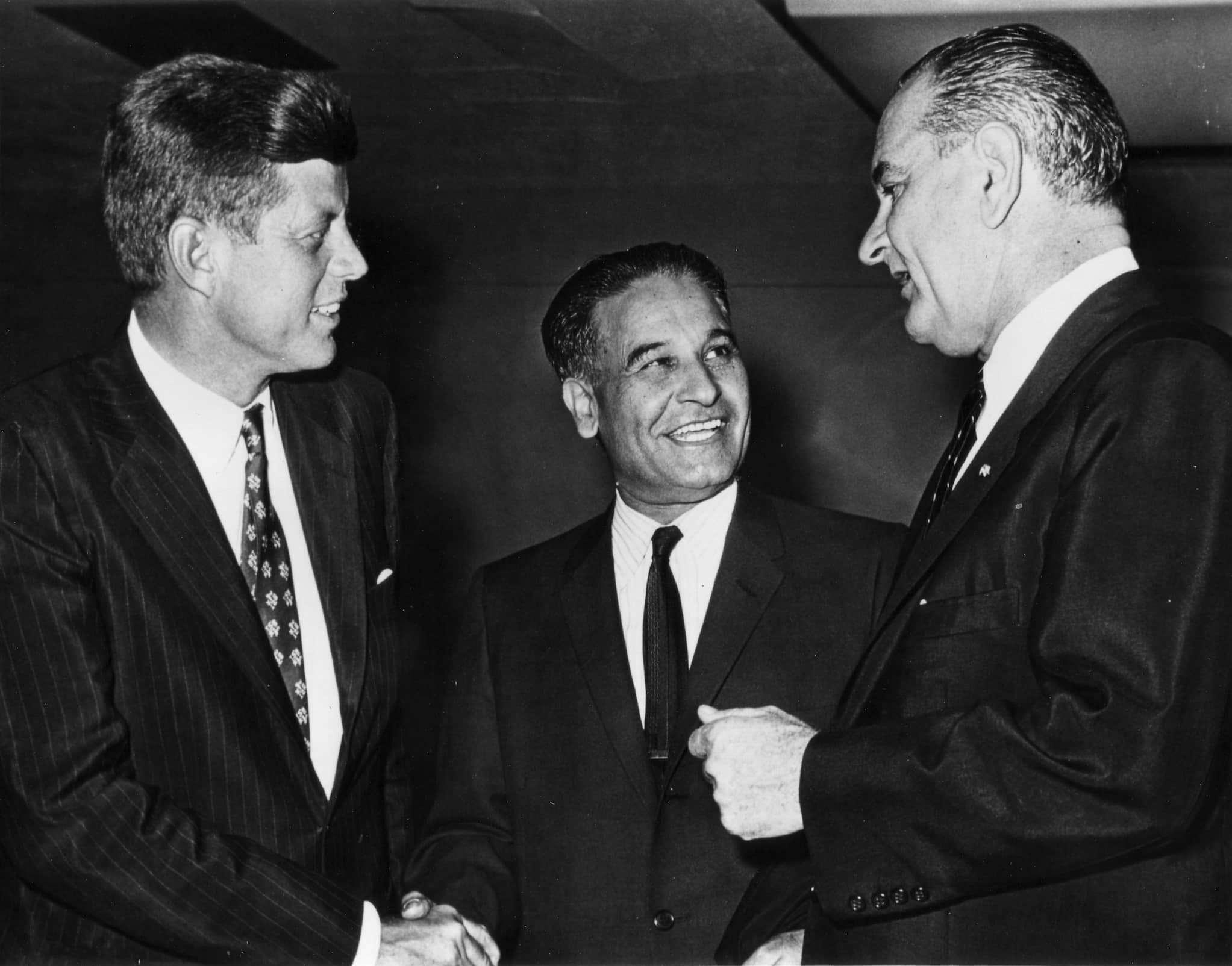 Indian American Dalip Singh Saund stands between John F. Kennedy and Lyndon Johnson. He shakes Kennedy's hand while looking at Johnson. All in suits.