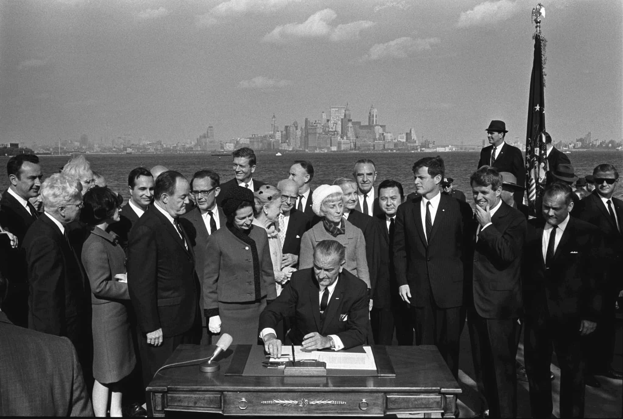 Seated at outdoor desk, President Lyndon B. Johnson signs the Hart-Celler Act of 1965 as government officials around him witness the signing.