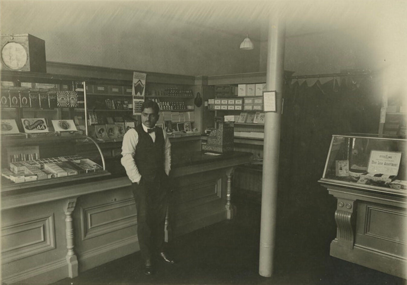 Indian Vaishno Das Bagai in a black vest, pants, bowtie, long sleeved collared shirt stands at the counter of his general store with hands in pockets.