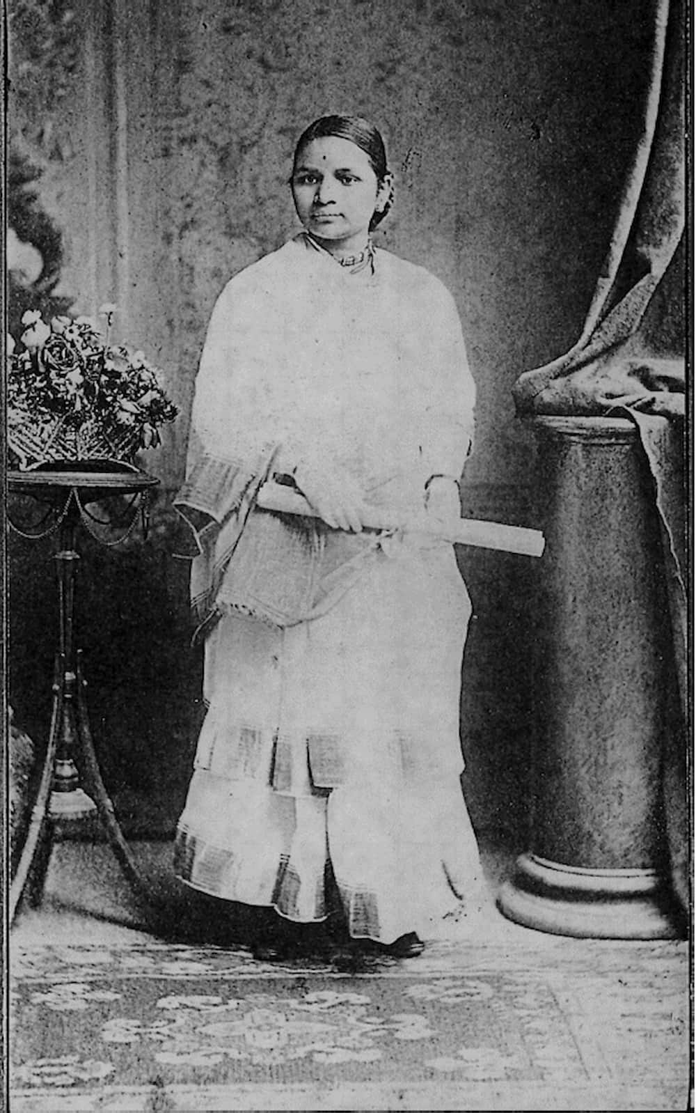 Indian Dr. Anandibai Joshee poses for a studio portrait dressed in saree and holds her diploma in her hands while standing on a rug.