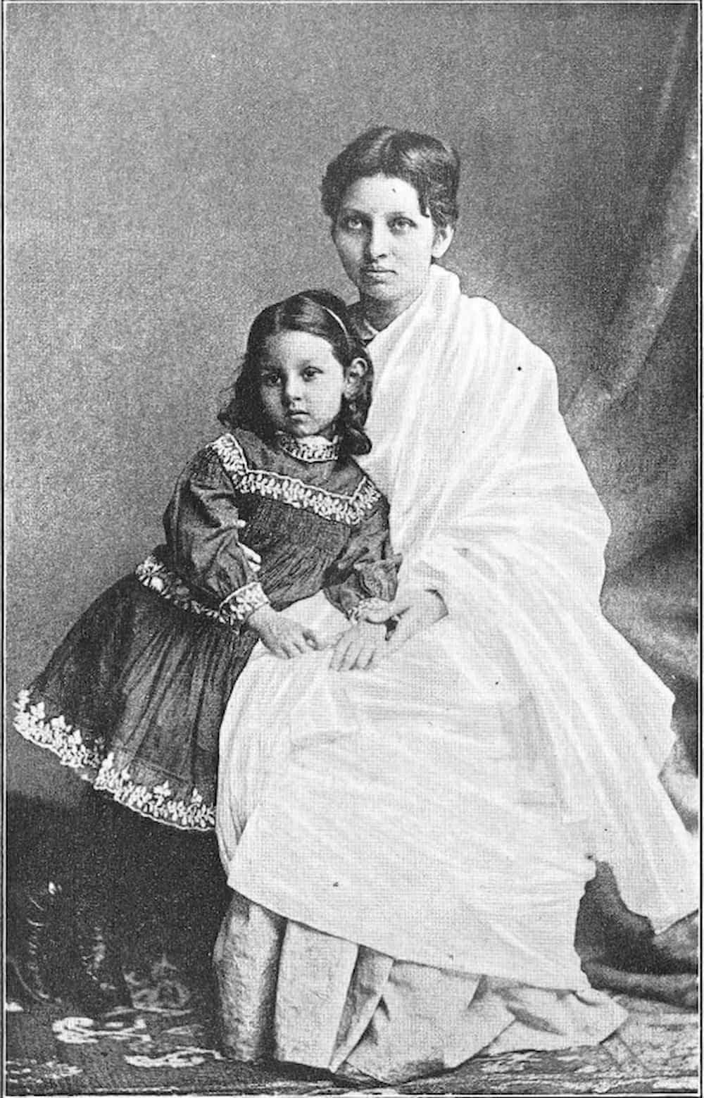 Portrait of Indian Pandita Ramabai. Dressed in saree, she sits while her young daughter in an ornately trimmed dress stands and leans against her.