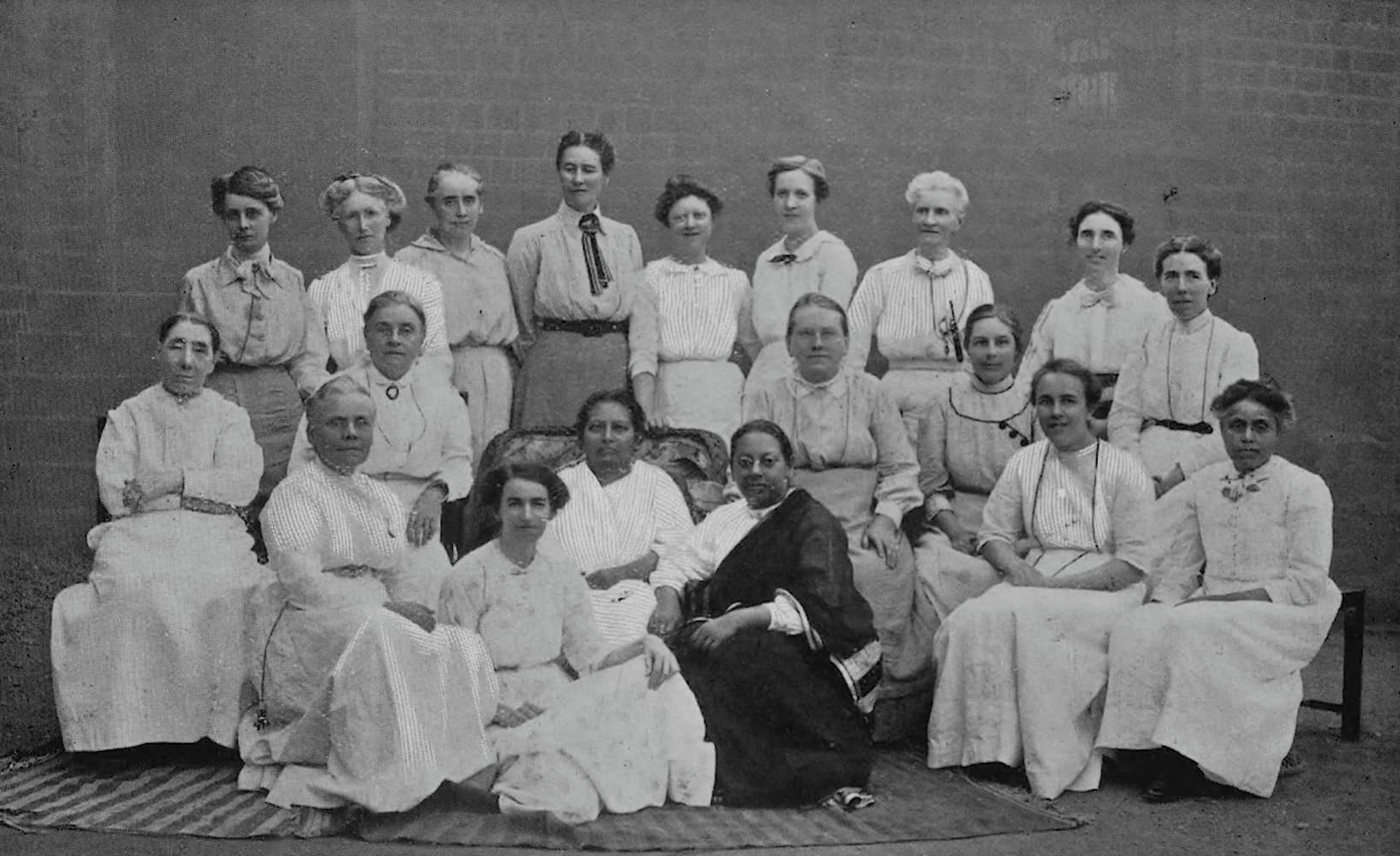 Pandita Ramabai and her daughter, both wearing saree, sit at center for formal portrait. Mukti Mission workers sit and stand beside them.
