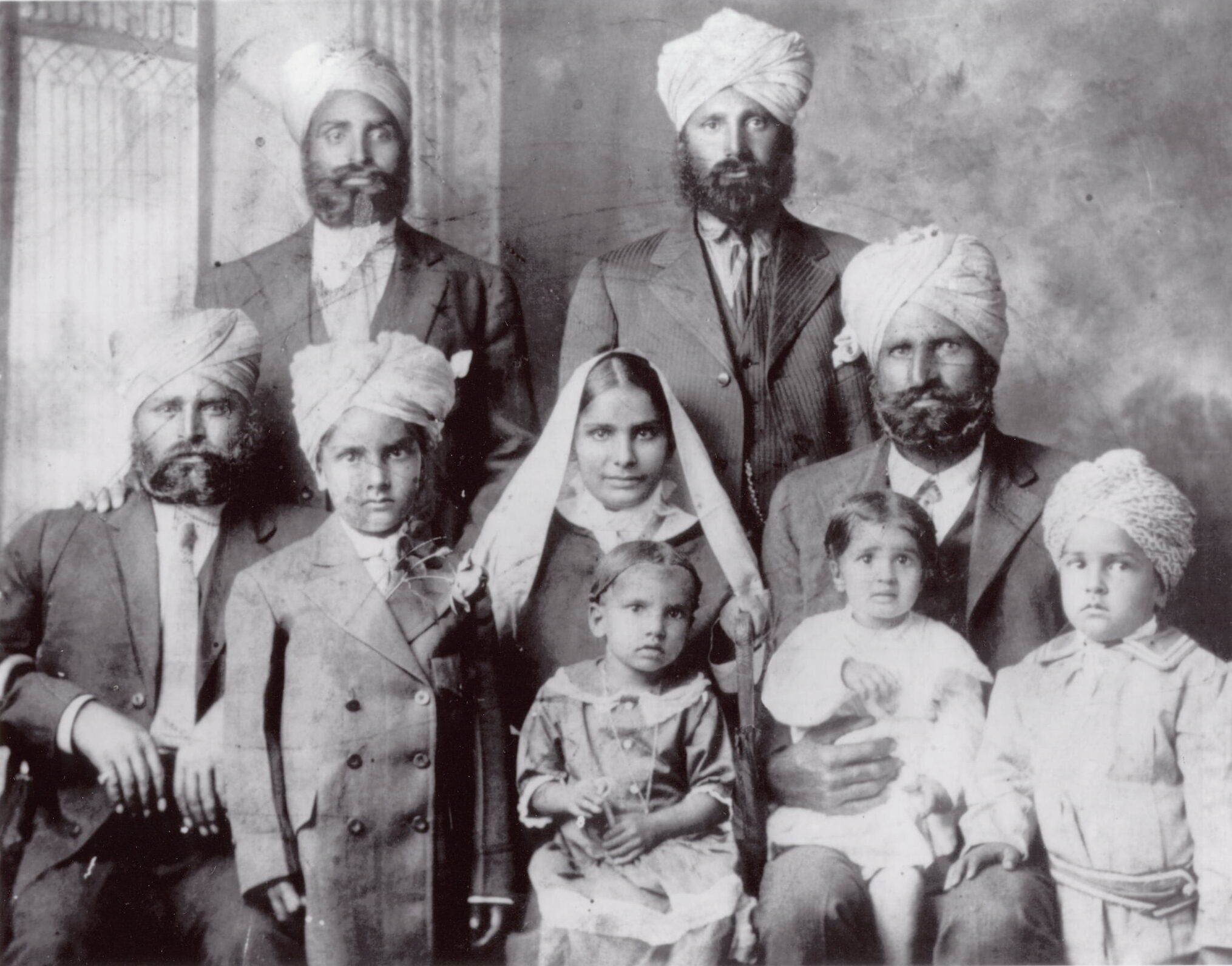 Family portrait of Indian American Kar Dhillon sitting on her father's lap. Six men and two boys wear turbans. A sibling sits on her mother's lap.