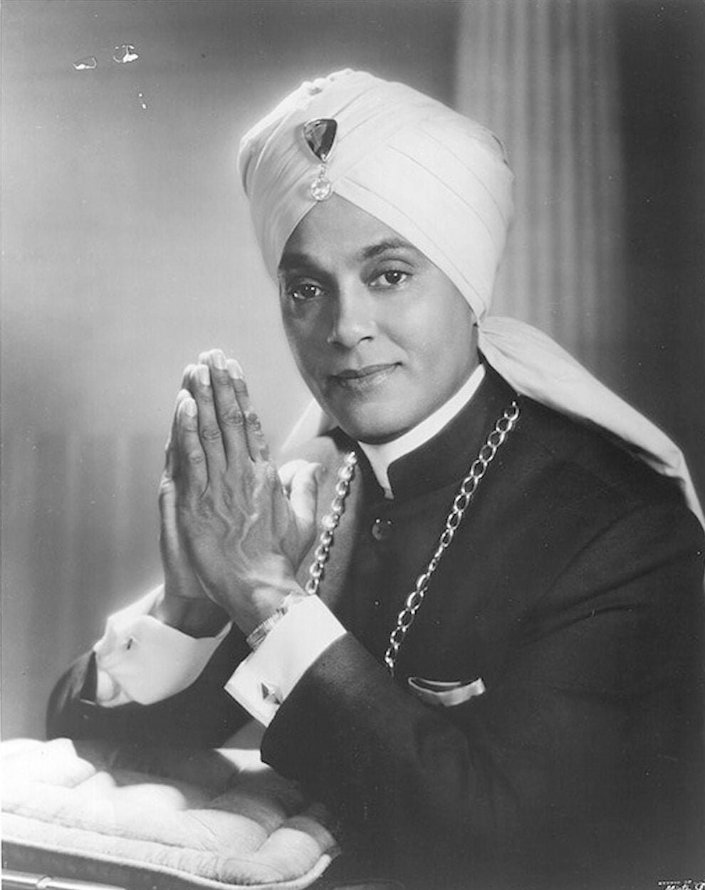 Pianist Korla Pandit poses for studio portrait. He holds his hands in prayer positions and wears a turban with a jewel at center.