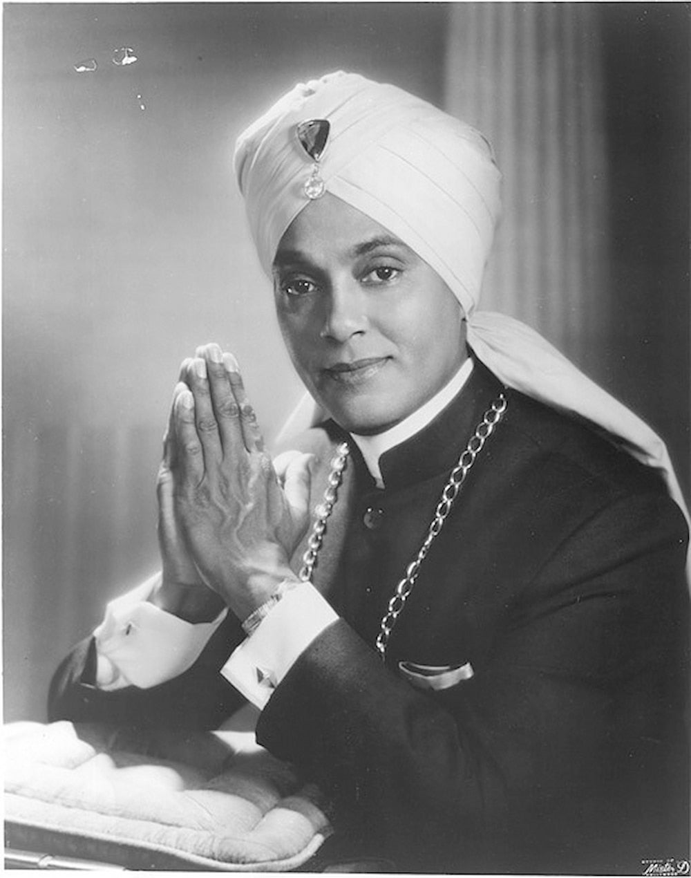 Pianist Korla Pandit poses for studio portrait. He holds his hands in prayer positions and wears a turban with a jewel at center.