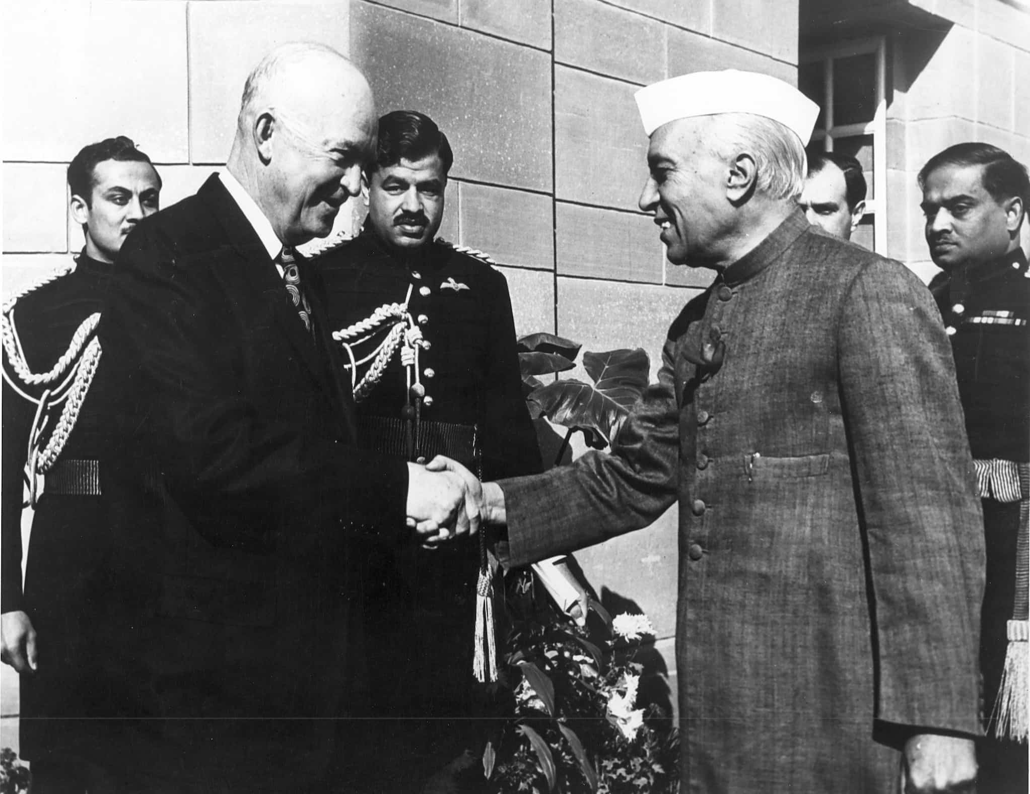 Indian Prime Minister Jawaharlal Nehru in a Gandhi cap and tailored coat shakes hands with President Eisenhower. Four Indian officers watch.