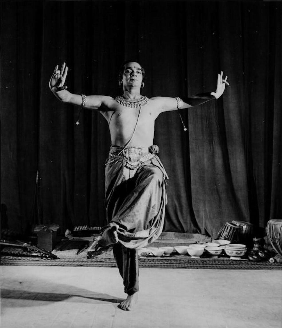 Indian Uday Shankar poses with his arms outstretched and hands in mudras. He stands with one leg raised and crossed in front of the other.