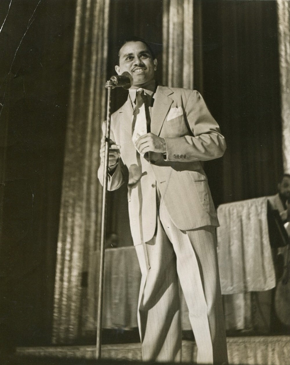 Indian musician Bardu Ali, wearing a suit and tie, stands in front of a microphone on a curtained stage.