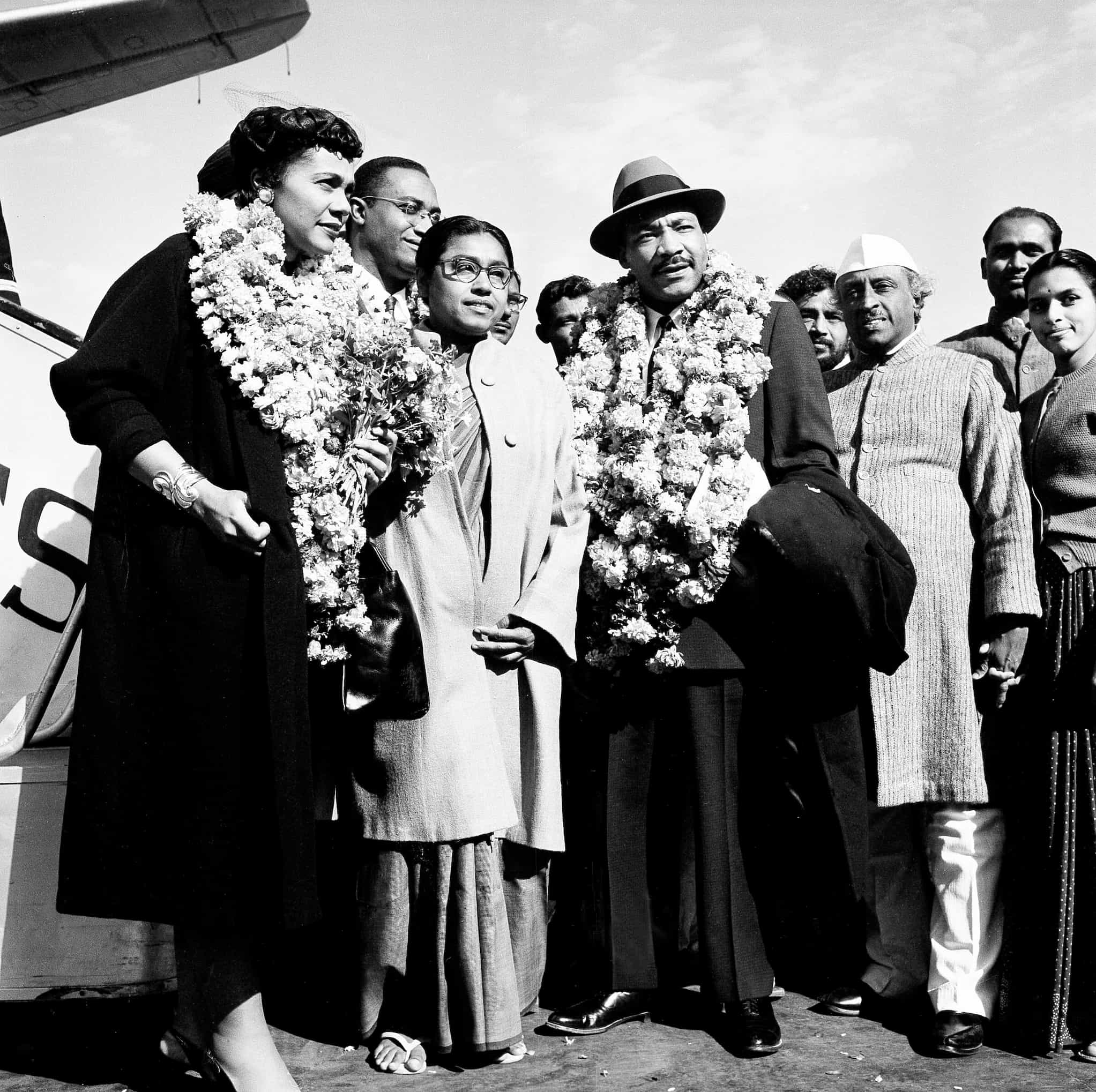 Dr. Martin Luther King, Jr. and Coretta Scott King, wearing floral garlands around their necks, stand with a group of Indian delegates on tarmac.