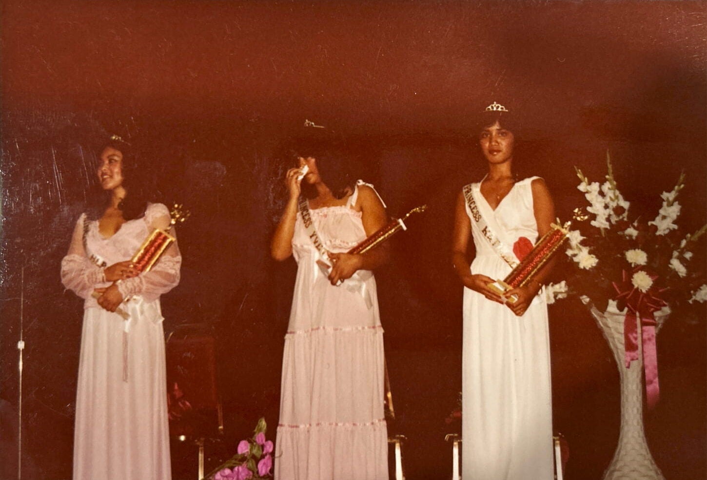 Three young women in formal gowns and tiaras hold trophies in their arms on a dark stage. Next to them is a standing floral arrangement.