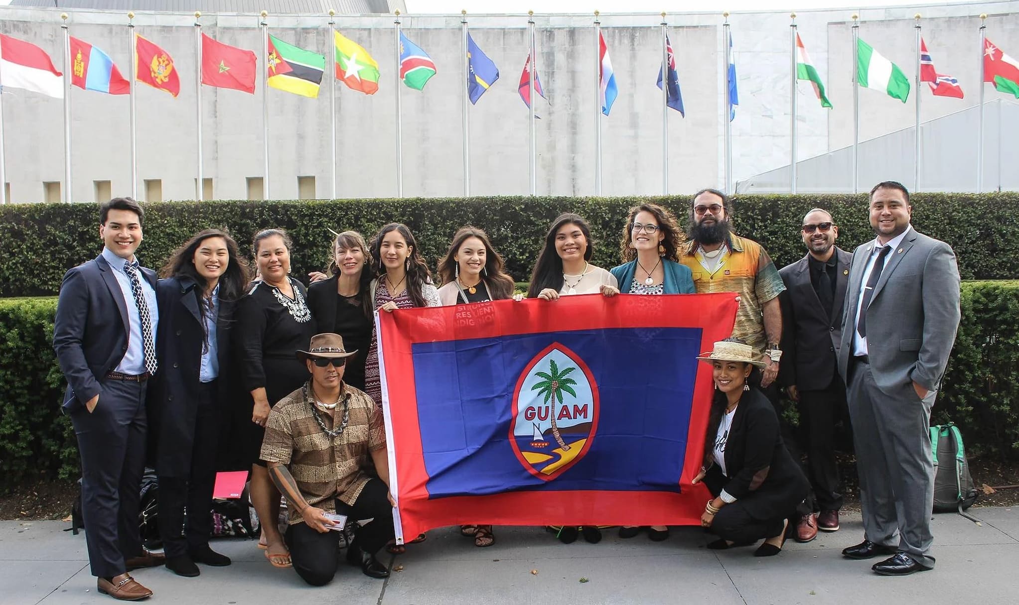 Thirteen CHamoru delegates pose for a picture in front of the flags at the United Nations' entrance. Together they hold up a large Guam flag.