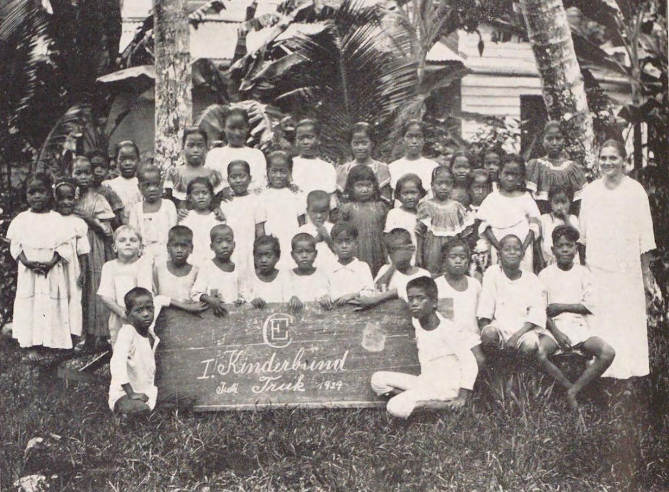Kindergarteners sit and stand on grassy lawn for outdoor photo. The thirty-six students gather around a large wooden sign on the ground.