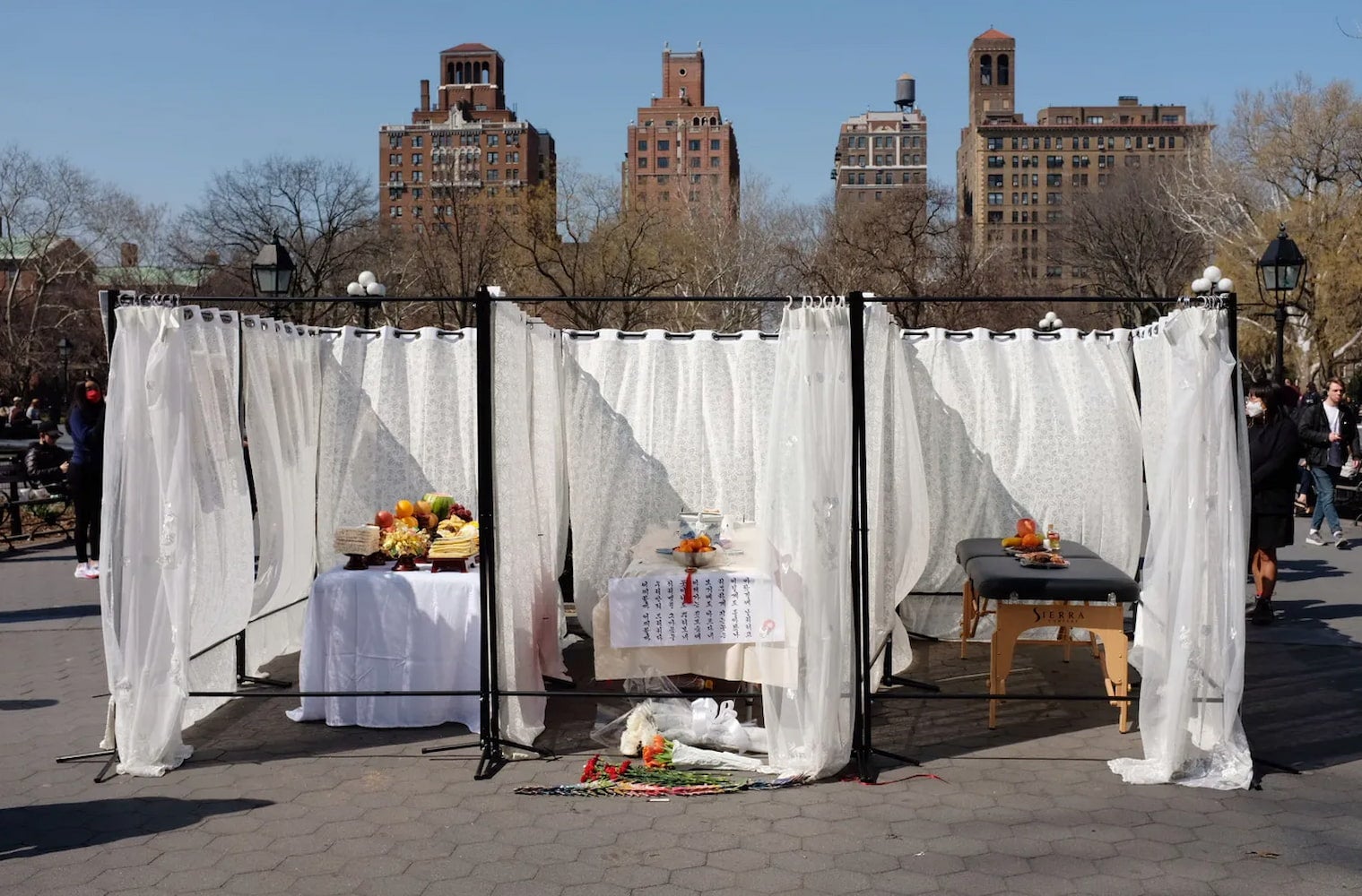 In a sunlit park, three massage tables separated by white curtains are used as memorial altars holding fruit and flower offerings.