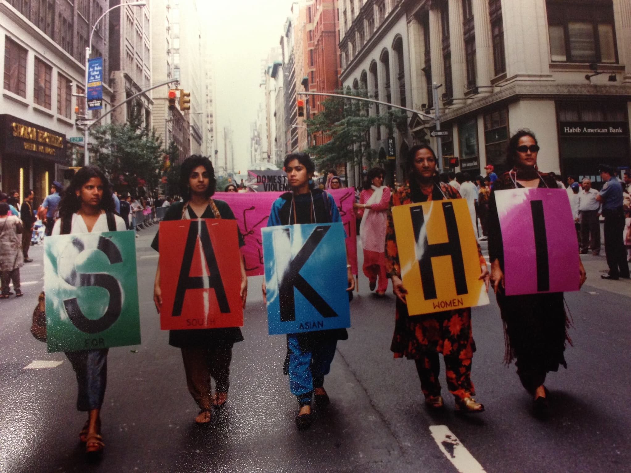 Five South Asian women march in a row on a street between tall buildings. They each hold multi-colored signs that spell out the word "Sakhi."